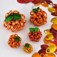 Five chainmaille pumpkins arranged on a light surface with autumn-colored scales scattered to the right. The pumpkins are different sizes and different shades of orange, with small green scales forming leaves at the top of each pumpkin.